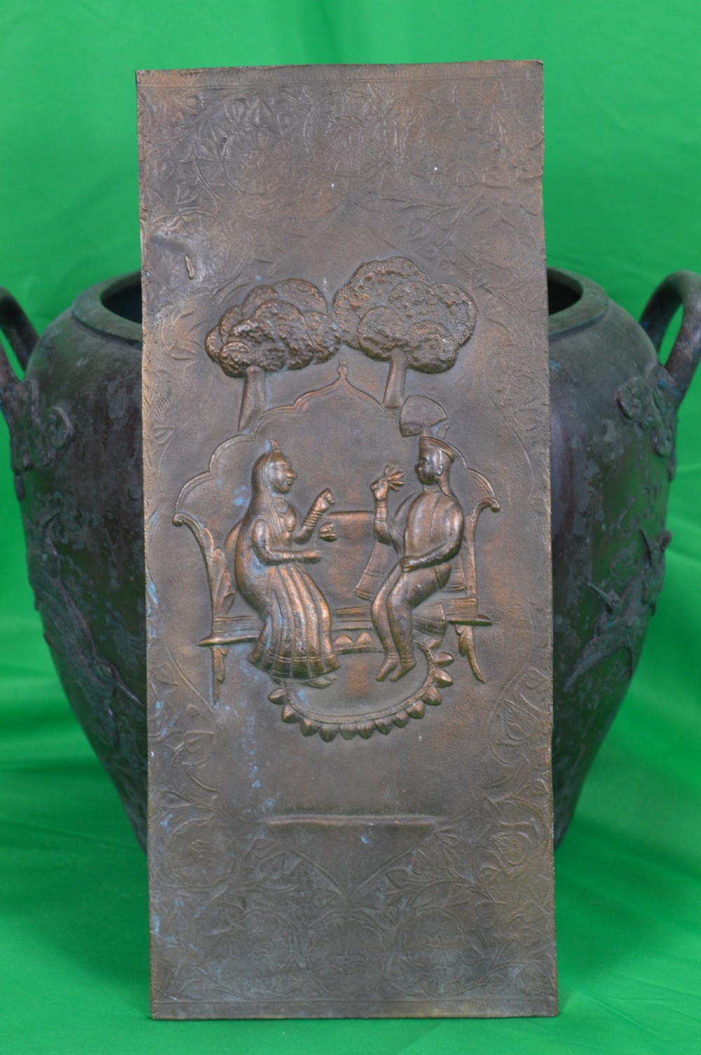 A rectangular antique bronze plaque featuring repousse metalwork depicting two seated figures under trees within a foliage border.
