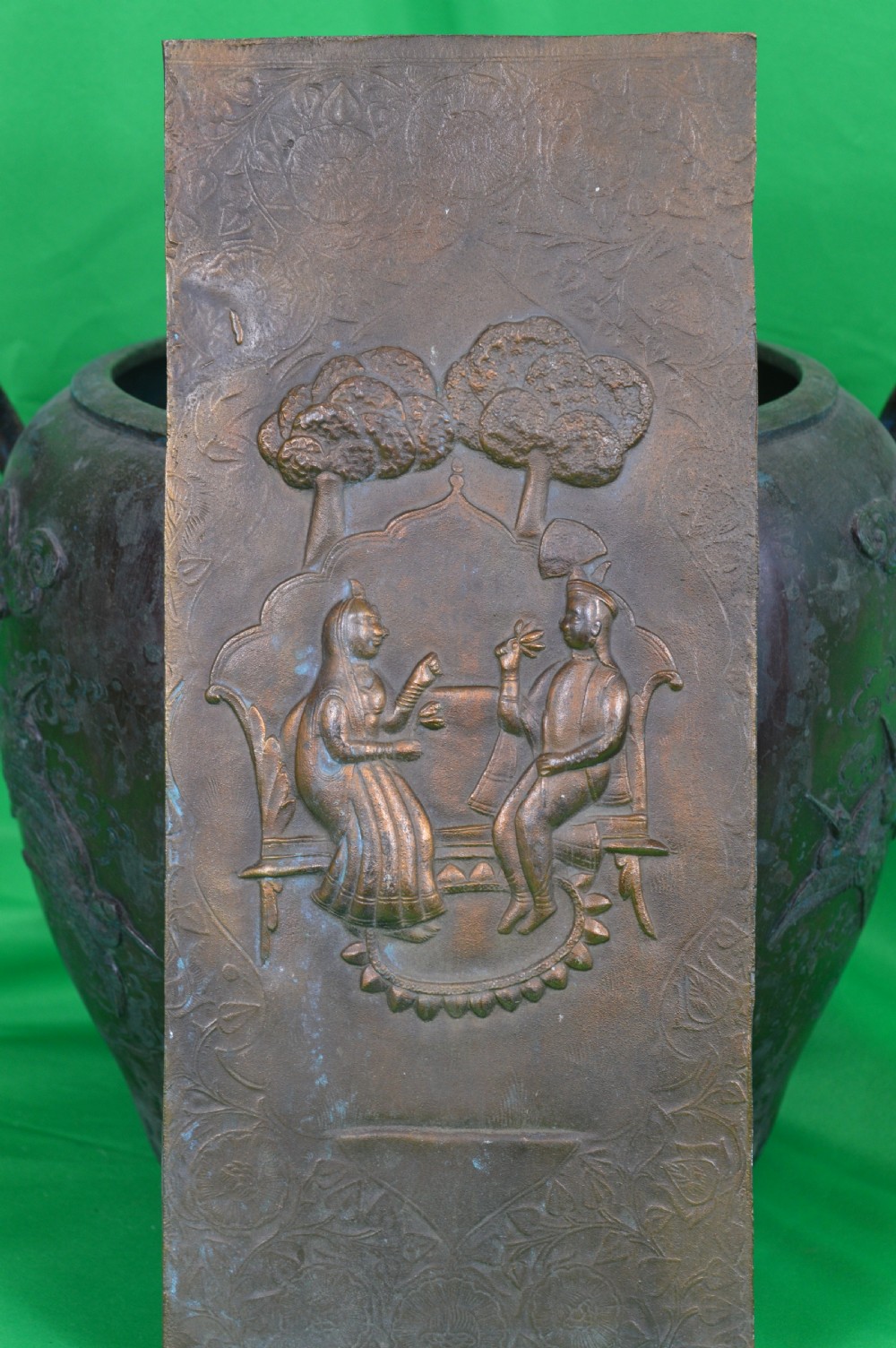 A rectangular antique bronze plaque featuring repousse metalwork depicting two seated figures under trees within a foliage border. View II
