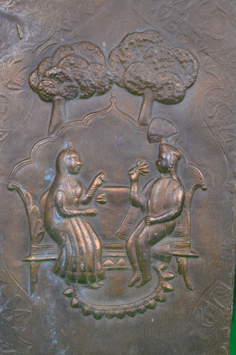 A rectangular antique bronze plaque featuring repousse metalwork depicting two seated figures under trees within a foliage border. View III