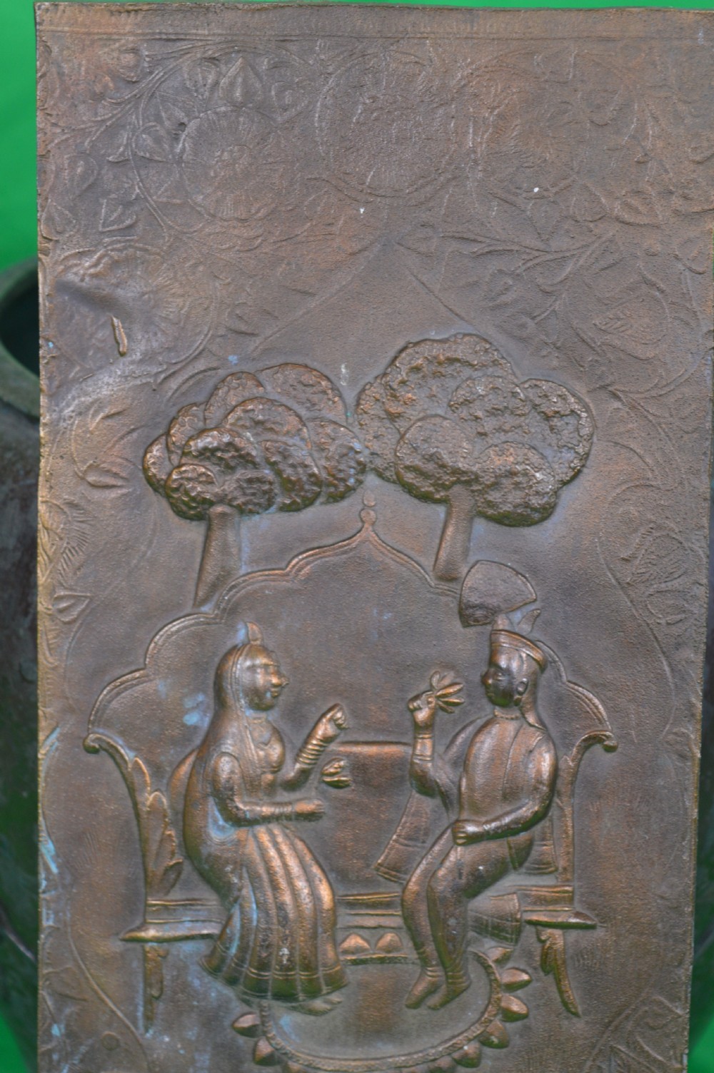 A rectangular antique bronze plaque featuring repousse metalwork depicting two seated figures under trees within a foliage border. View IV