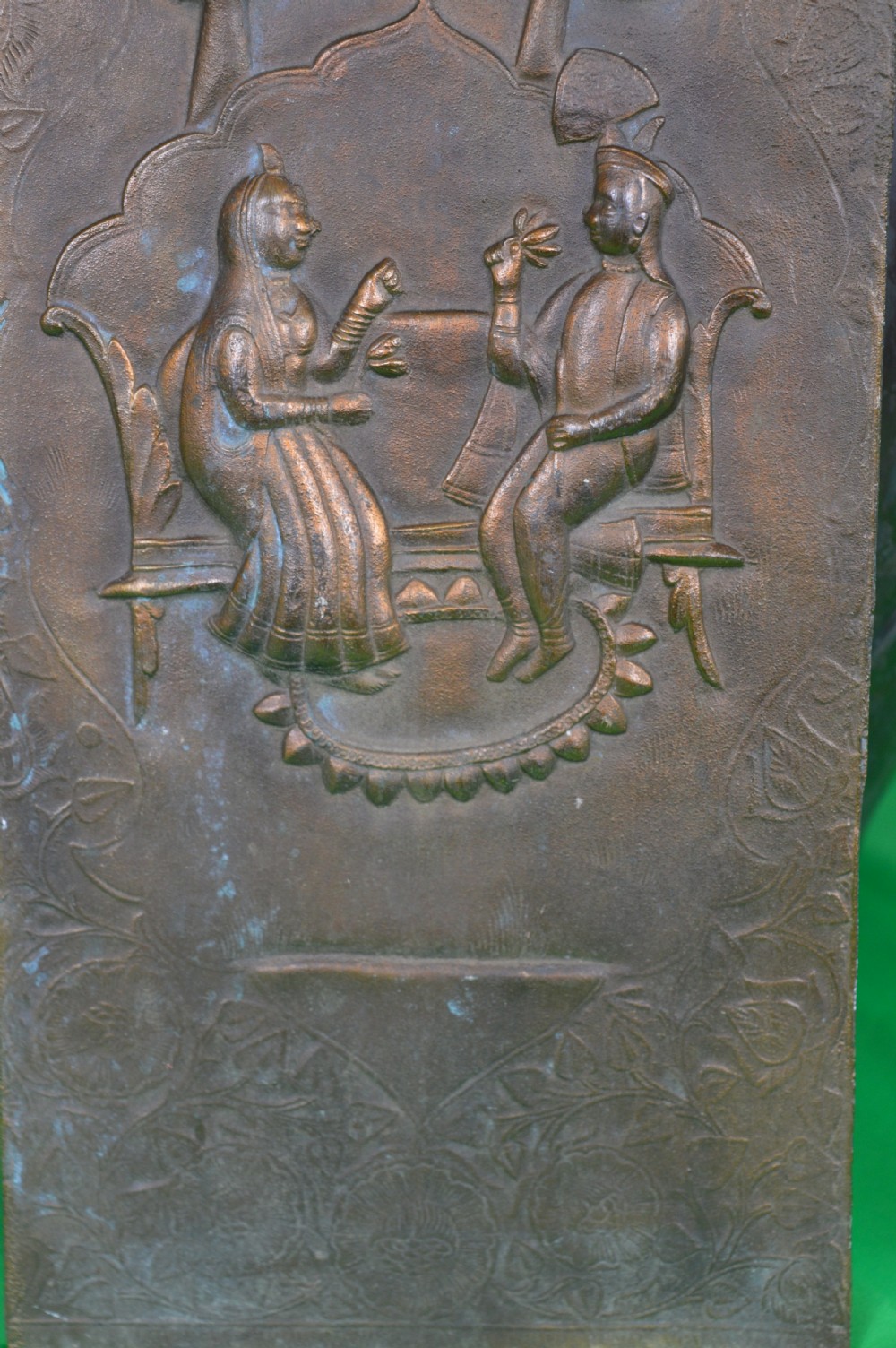 A rectangular antique bronze plaque featuring repousse metalwork depicting two seated figures under trees within a foliage border. View V