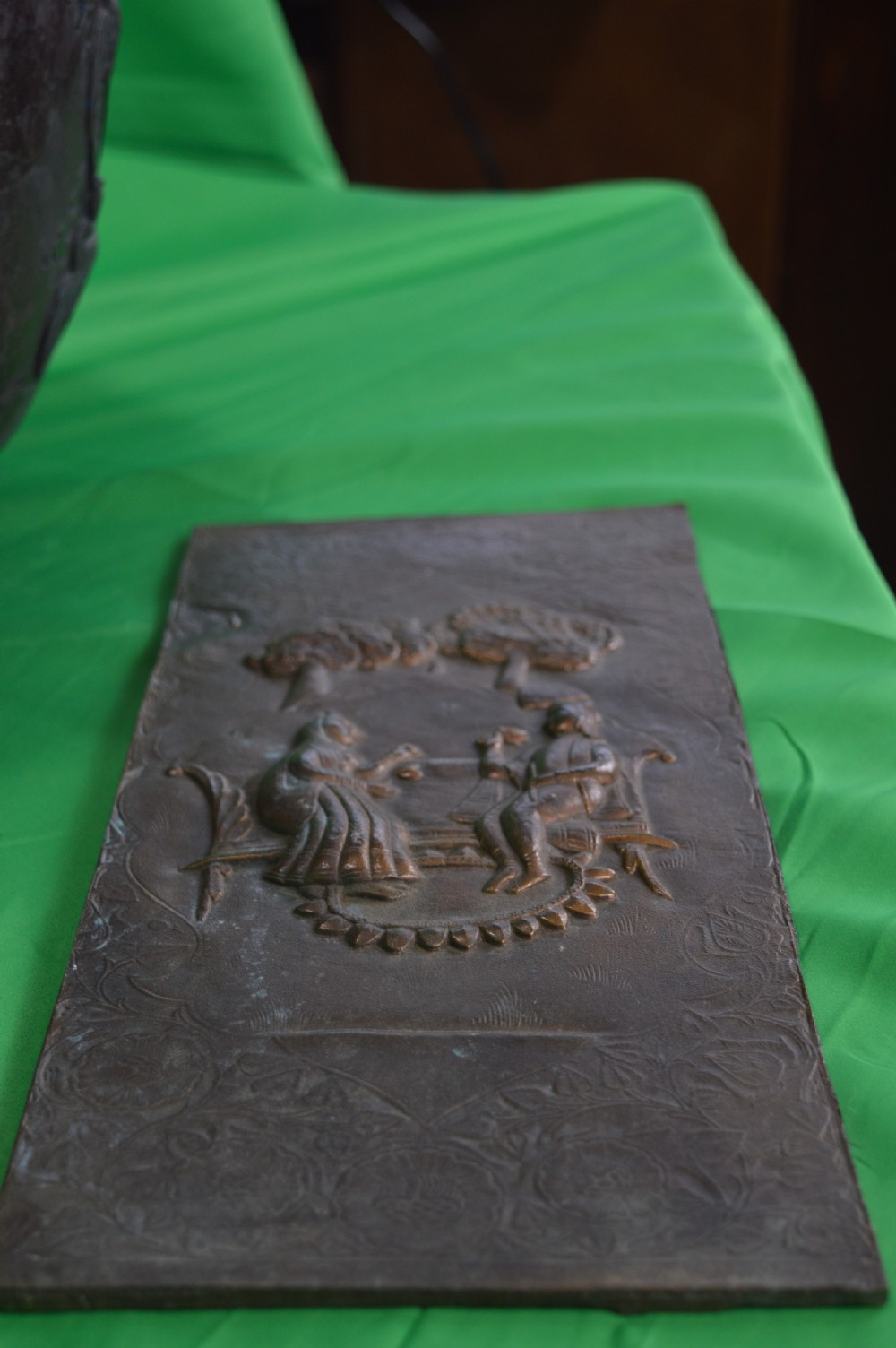 A rectangular antique bronze plaque featuring repousse metalwork depicting two seated figures under trees within a foliage border. View VIII