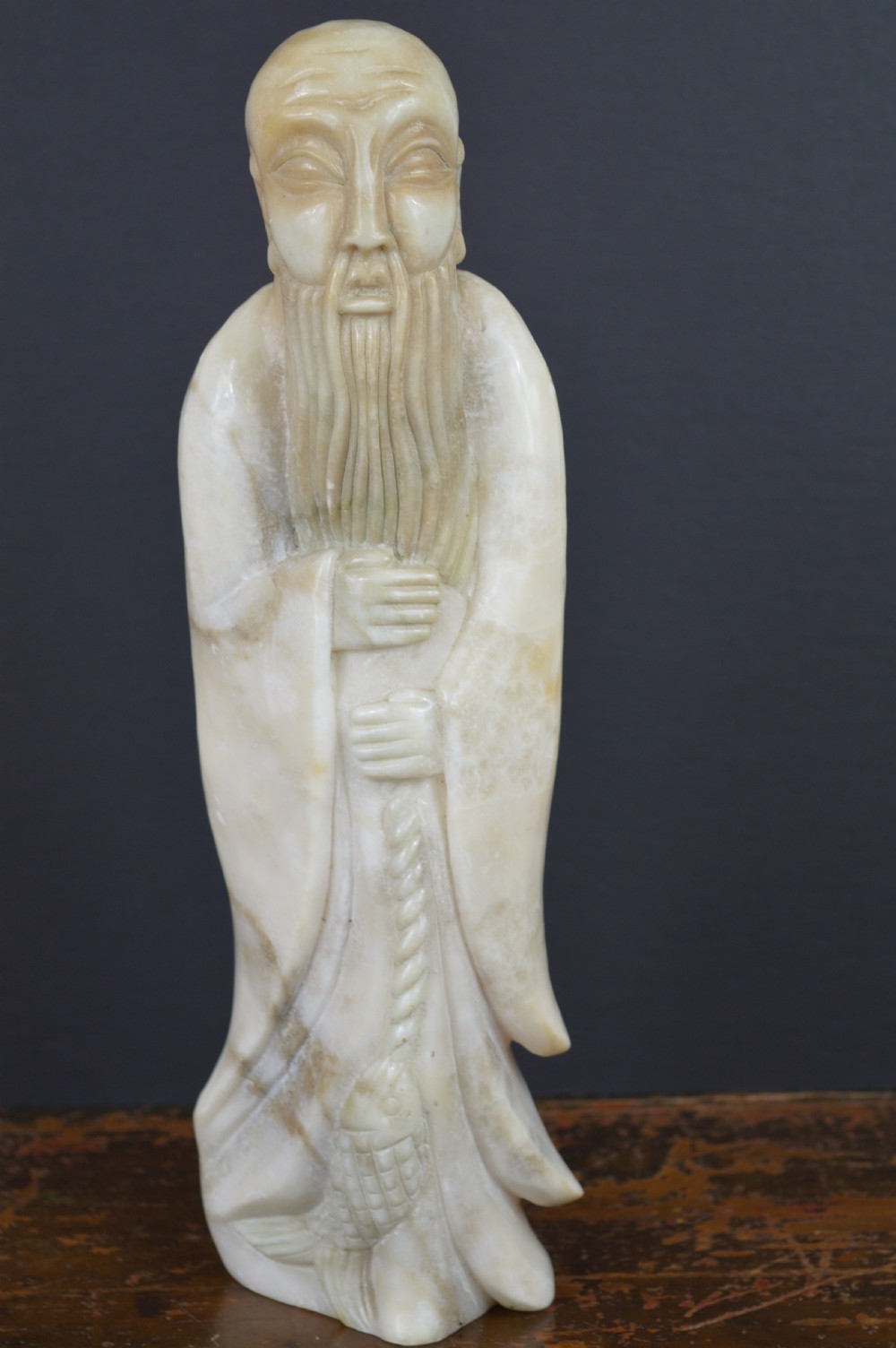 A cream-coloured carved hardstone sculpture depicting a standing bearded Chinese sage in long flowing robes holding a fish on a cord.