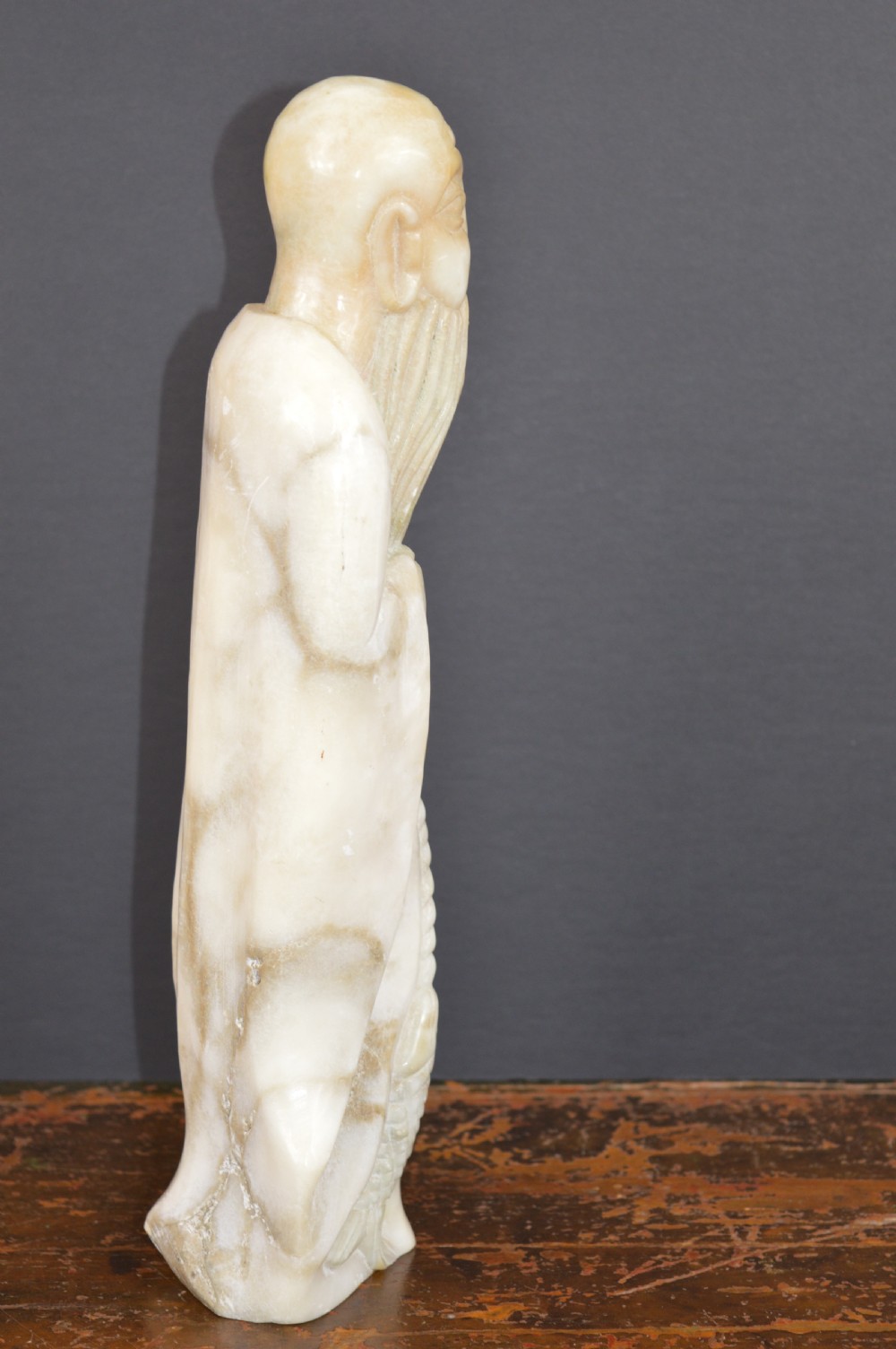 A cream-coloured carved hardstone sculpture depicting a standing bearded Chinese sage in long flowing robes holding a fish on a cord. View II