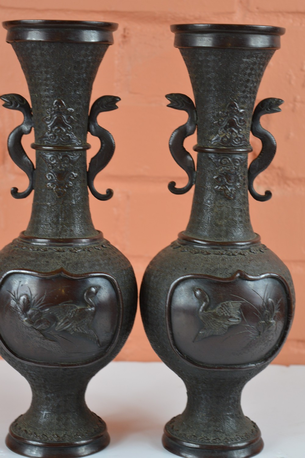 A pair of dark brown patinated Japanese bronze vases featuring dragon-form handles and relief panels depicting birds amongst aquatic foliage.