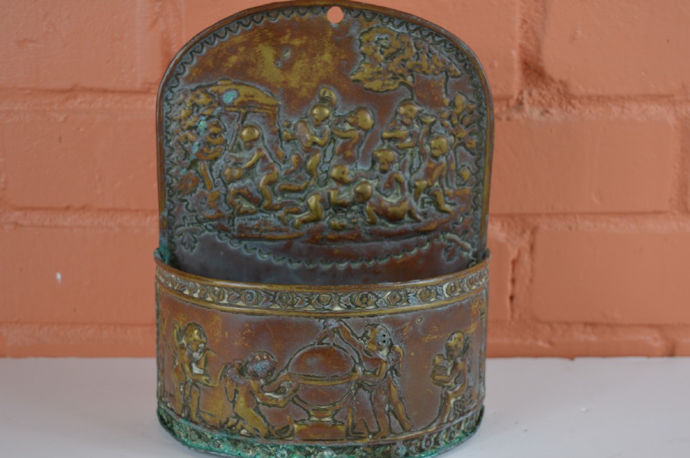 An antique embossed brass hanging spill box featuring detailed cherub motifs in repousse relief and an arched mounting backplate.
