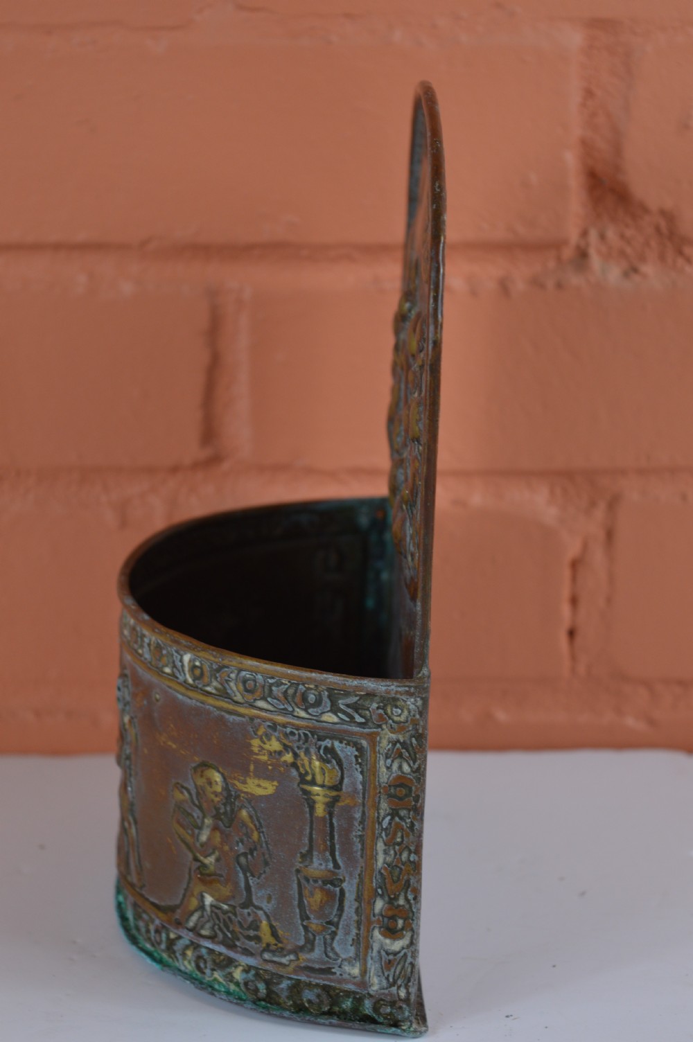 An antique embossed brass hanging spill box featuring detailed cherub motifs in repousse relief and an arched mounting backplate. View V