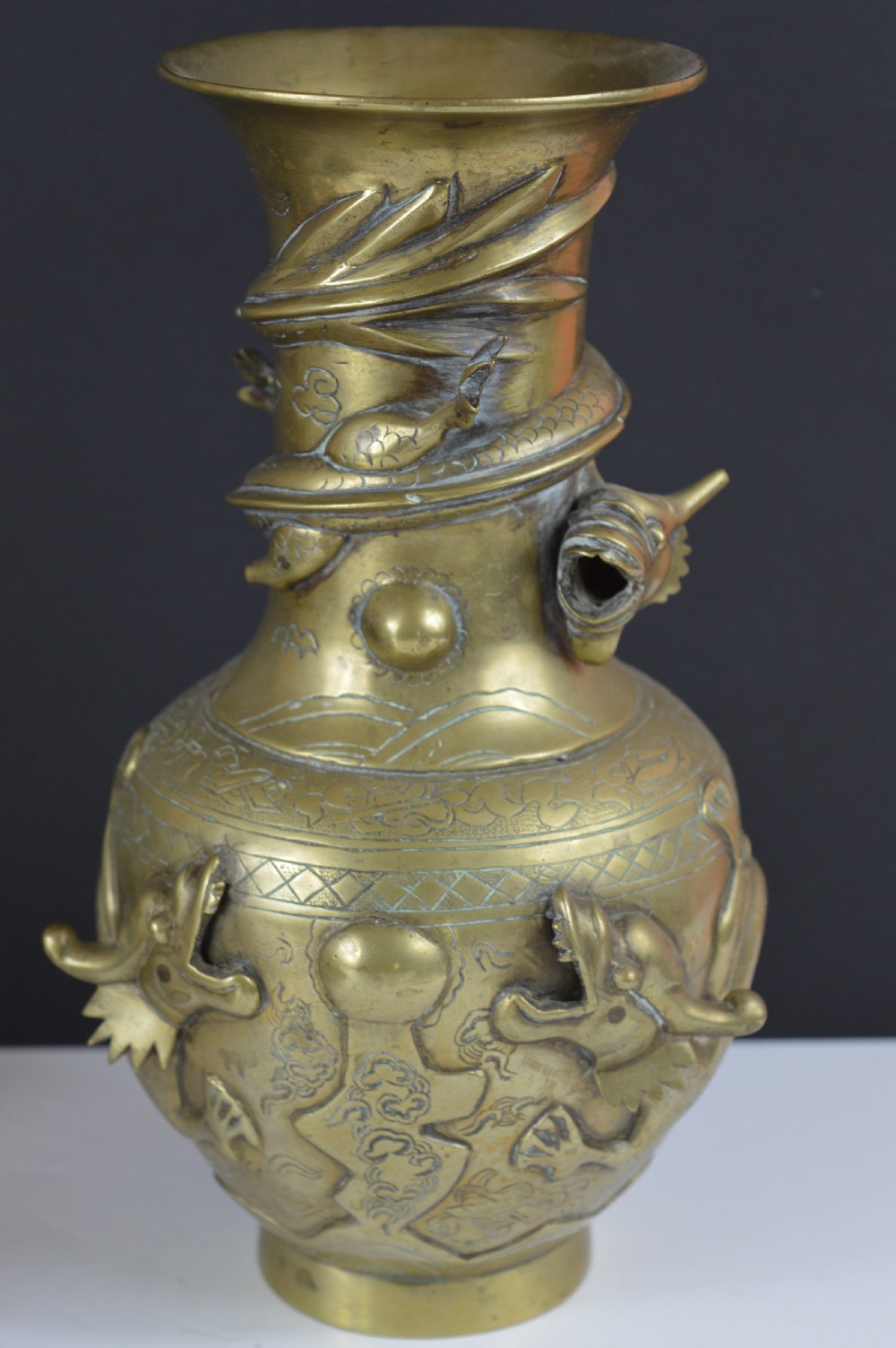 An antique brass baluster vase decorated with high-relief coiled dragons around the neck and body alongside engraved cloud patterns.