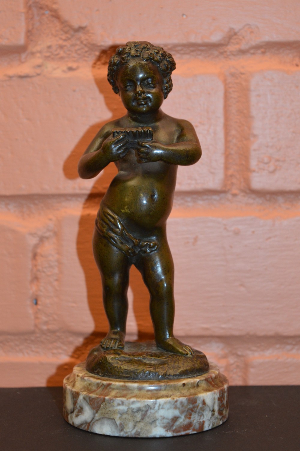 A dark bronze statue of a cherub playing pan pipes, mounted on a round veined marble base.