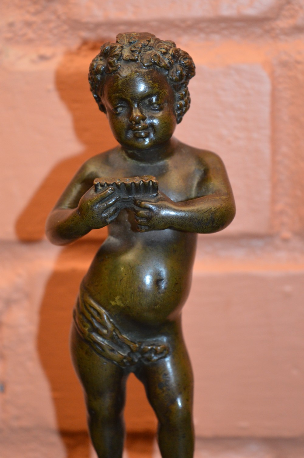 A dark bronze statue of a cherub playing pan pipes, mounted on a round veined marble base. View II