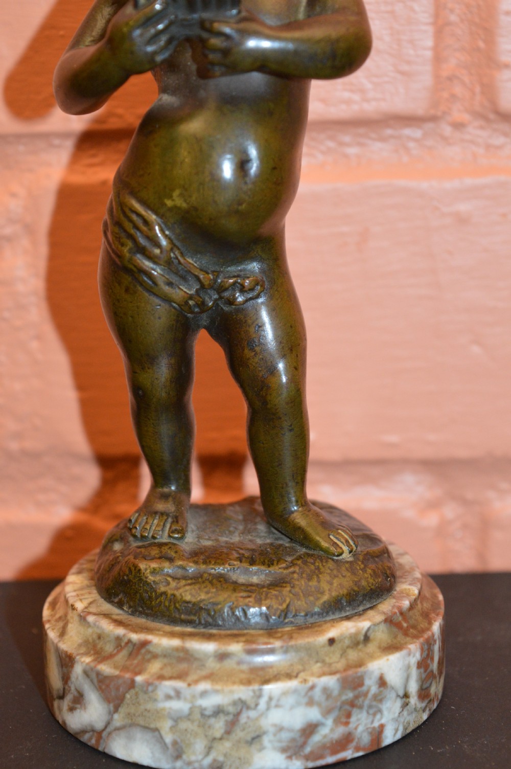 A dark bronze statue of a cherub playing pan pipes, mounted on a round veined marble base. View III