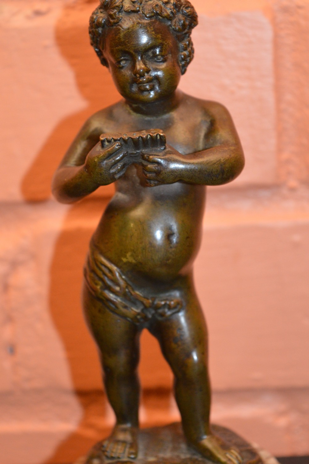 A dark bronze statue of a cherub playing pan pipes, mounted on a round veined marble base. View IV