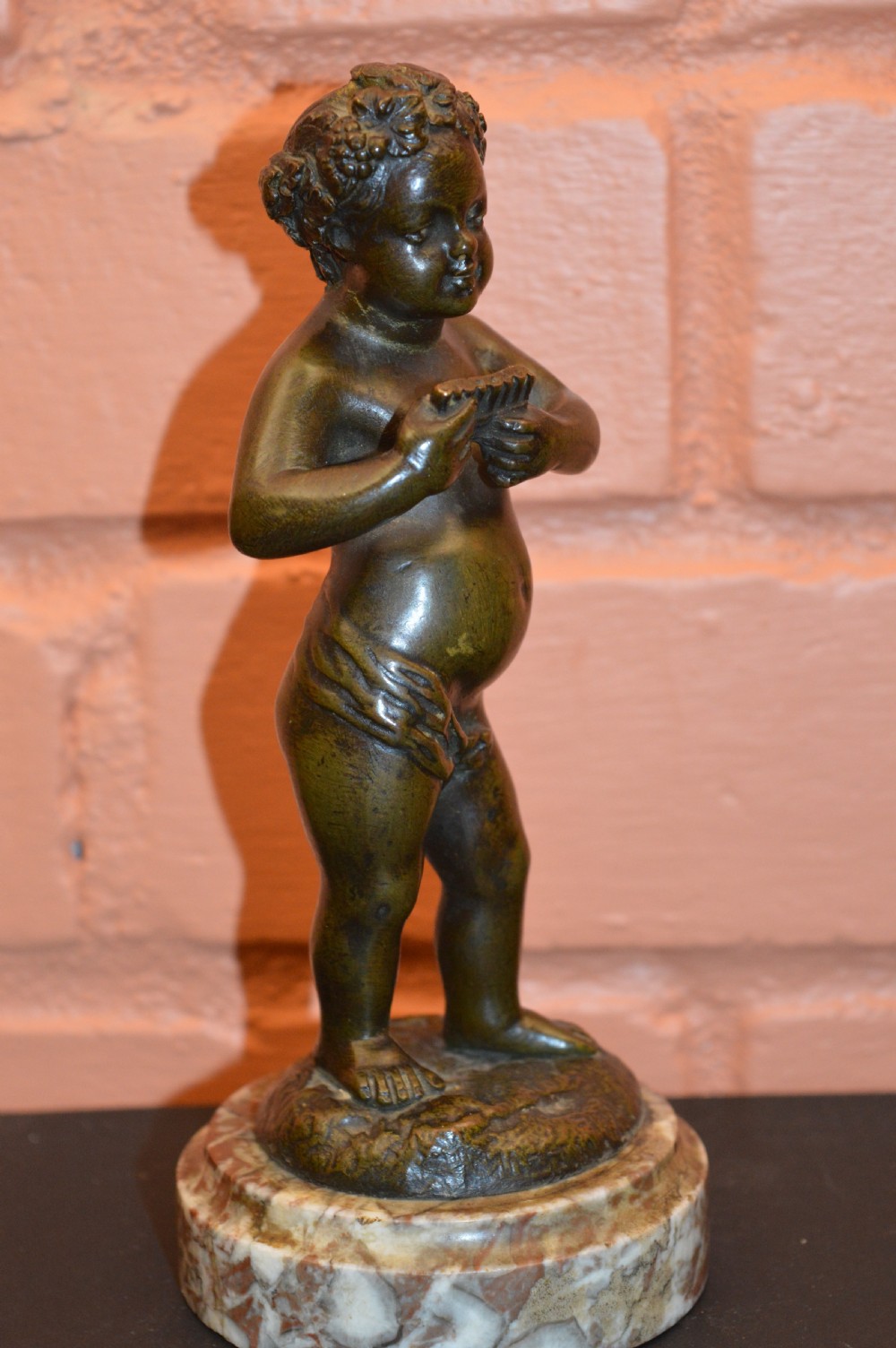 A dark bronze statue of a cherub playing pan pipes, mounted on a round veined marble base. View V