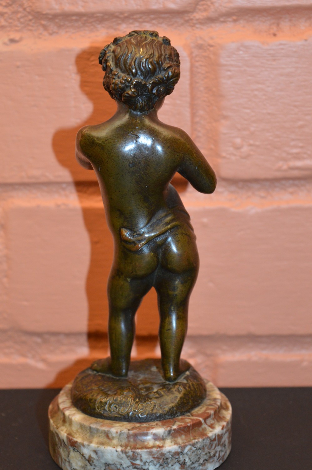 A dark bronze statue of a cherub playing pan pipes, mounted on a round veined marble base. View VI