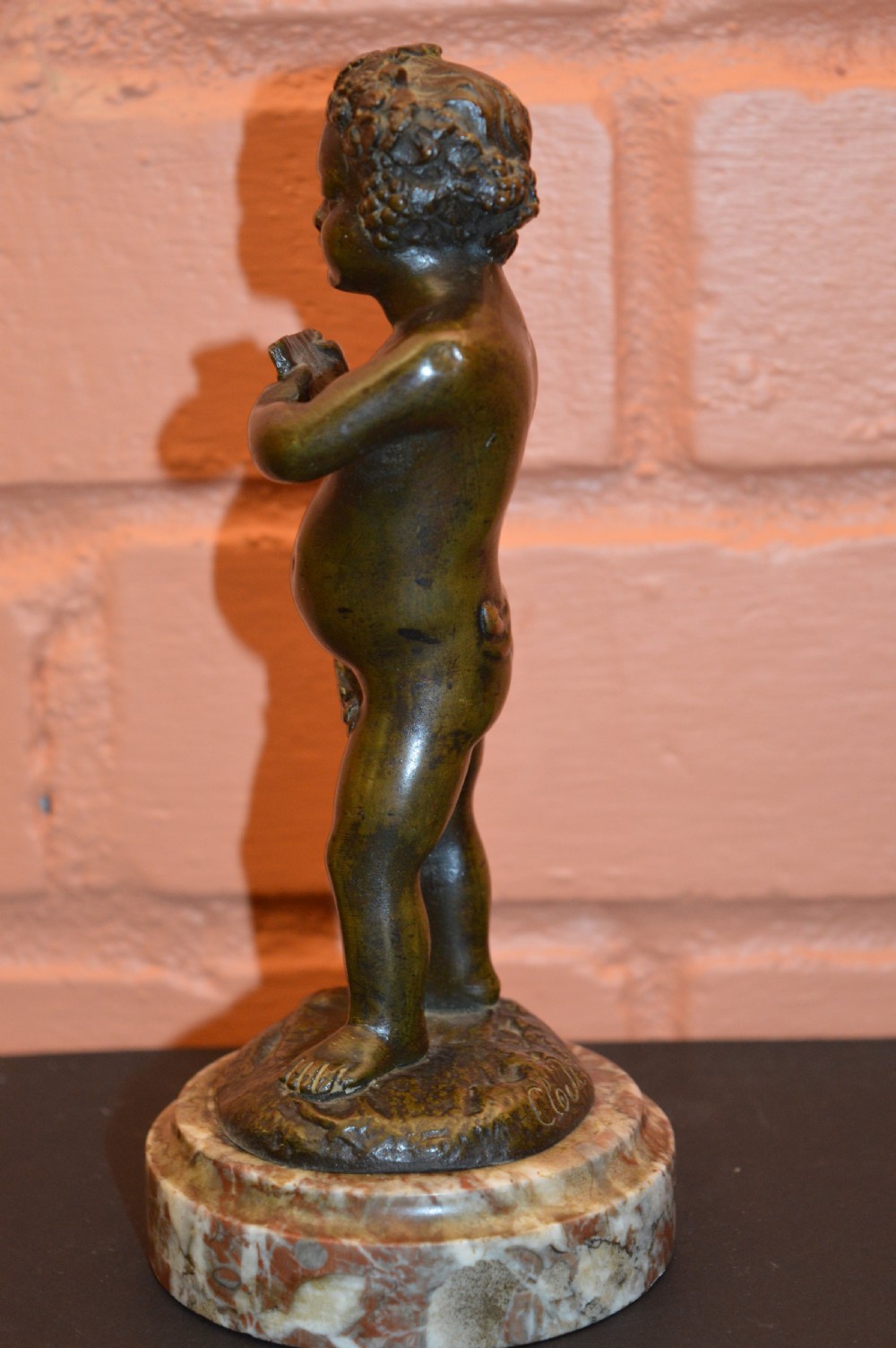 A dark bronze statue of a cherub playing pan pipes, mounted on a round veined marble base. View VII