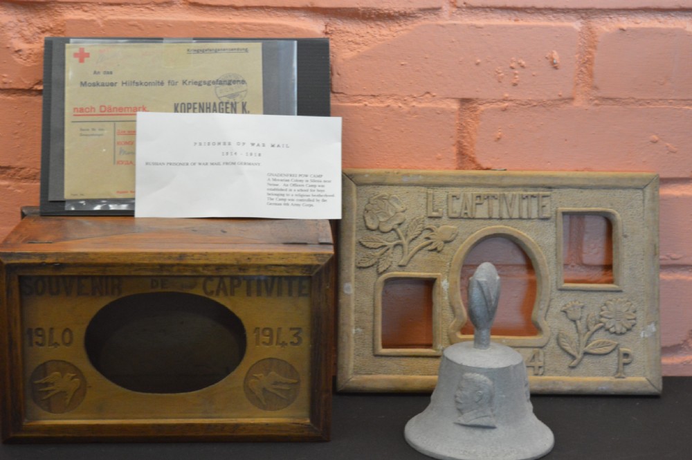 A four-piece collection of prisoner of war memorabilia including a carved wooden box, a wooden frame, a metal bell, and framed wartime documents.
