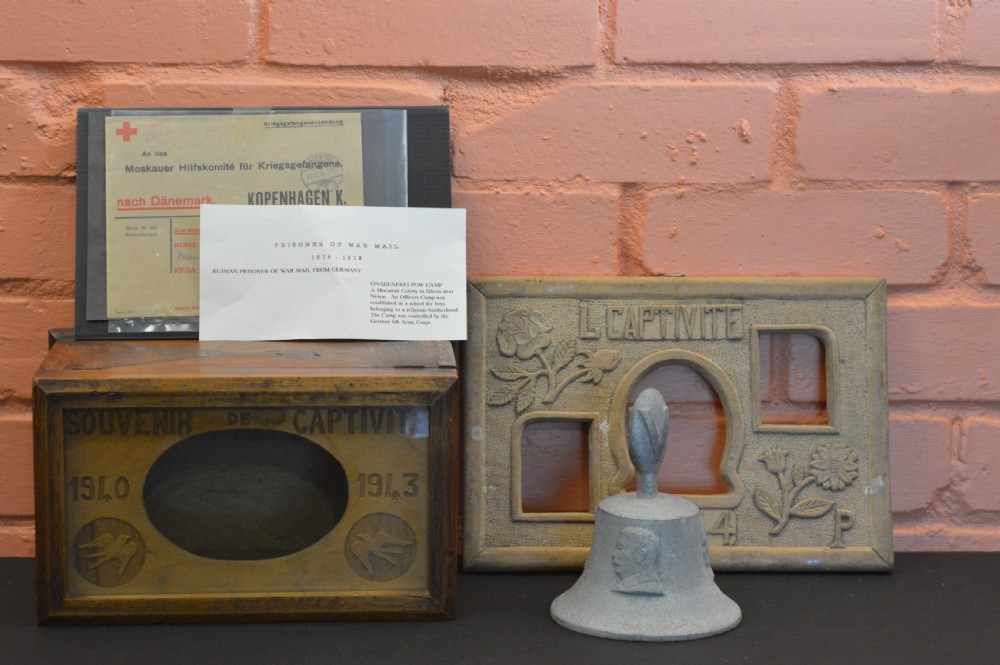 A four-piece collection of prisoner of war memorabilia including a carved wooden box, a wooden frame, a metal bell, and framed wartime documents. View II
