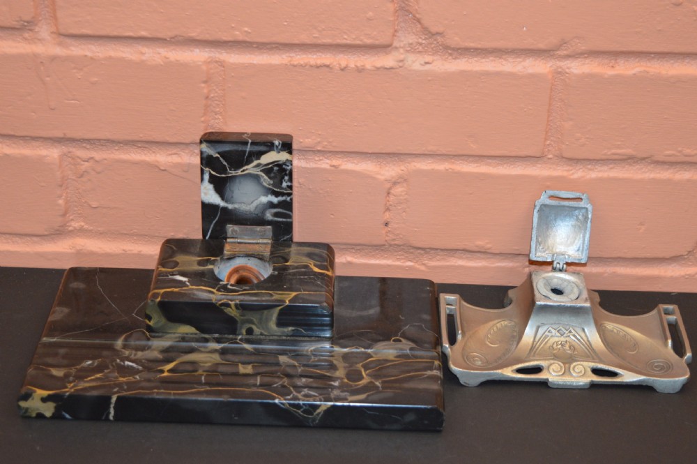 Two antique desk inkstands, one composed of black and gold veined marble with a built-in pen groove, and the second crafted from silver-toned metal with relief detailing.
