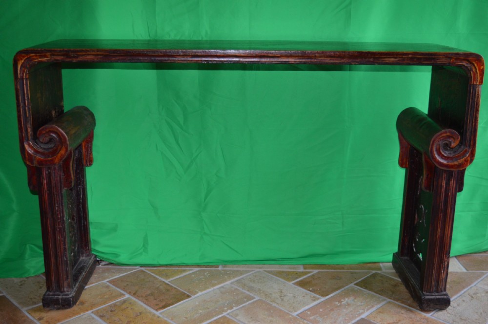 A long dark hardwood Tibetan altar table featuring a flat rectangular top supported by upright legs with prominent carved scroll terminals.