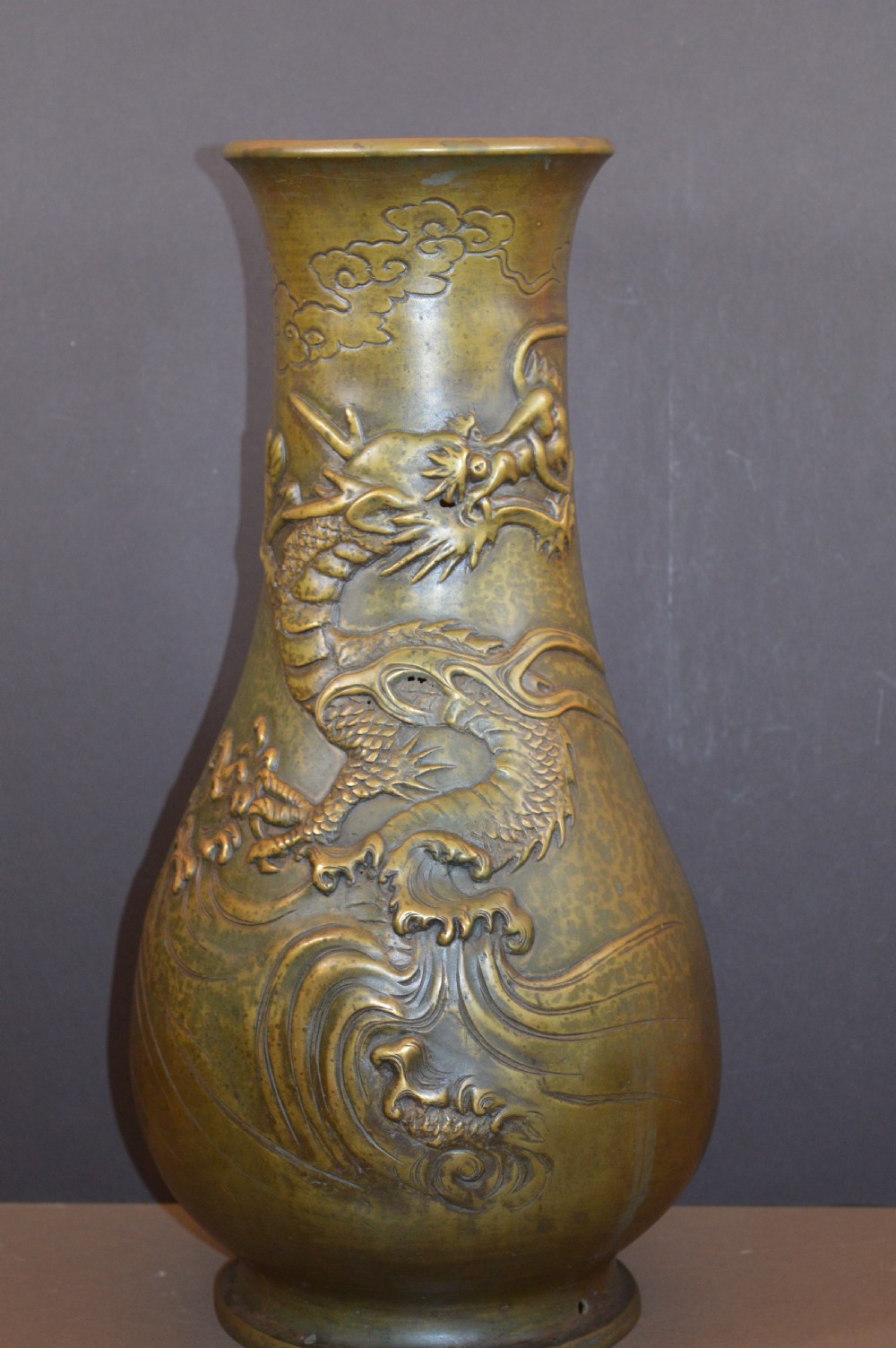 A tall baluster-shaped bronze vase featuring a high-relief carved dragon emerging from swirling waves and cloud motifs.