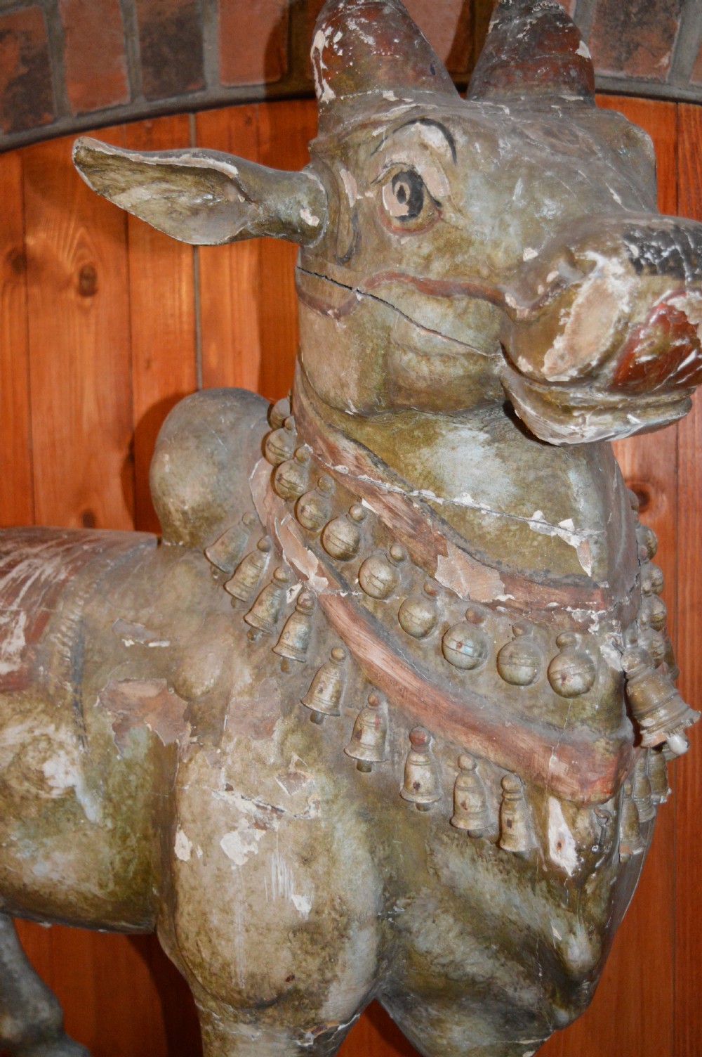 A large antique carved wooden Nandi bull statue featuring worn polychrome paint and decorative carved bells around its neck. View XII