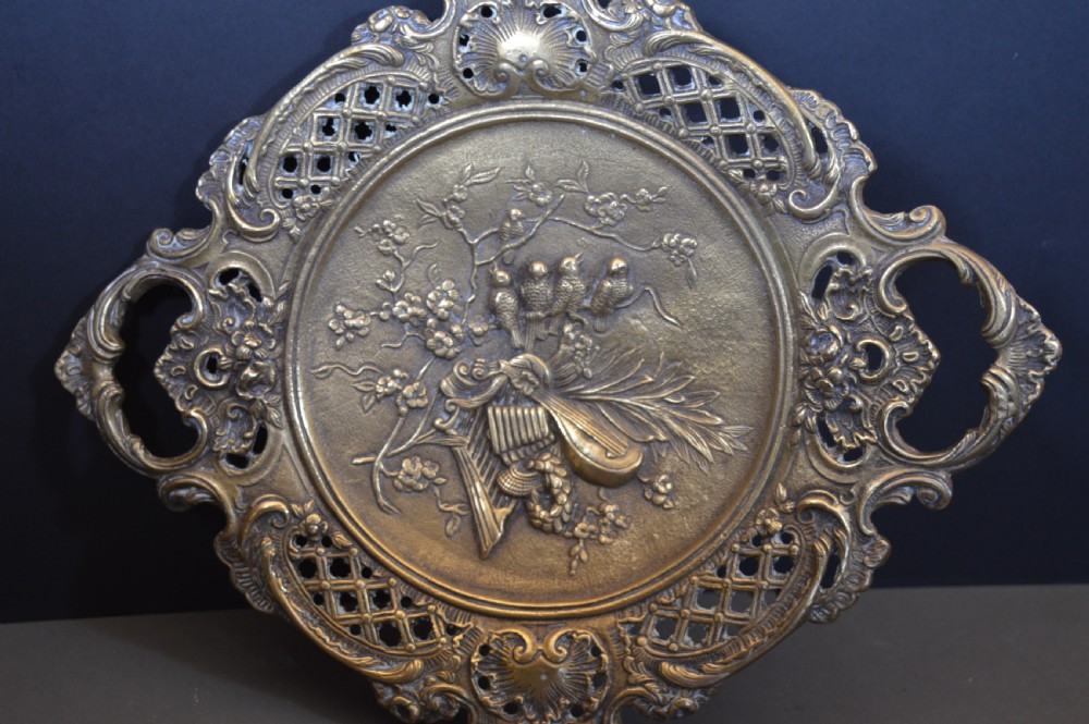 A cast bronze decorative centrepiece plate featuring a high-relief central scene with birds and musical instruments surrounded by an openwork lattice border and scroll handles.