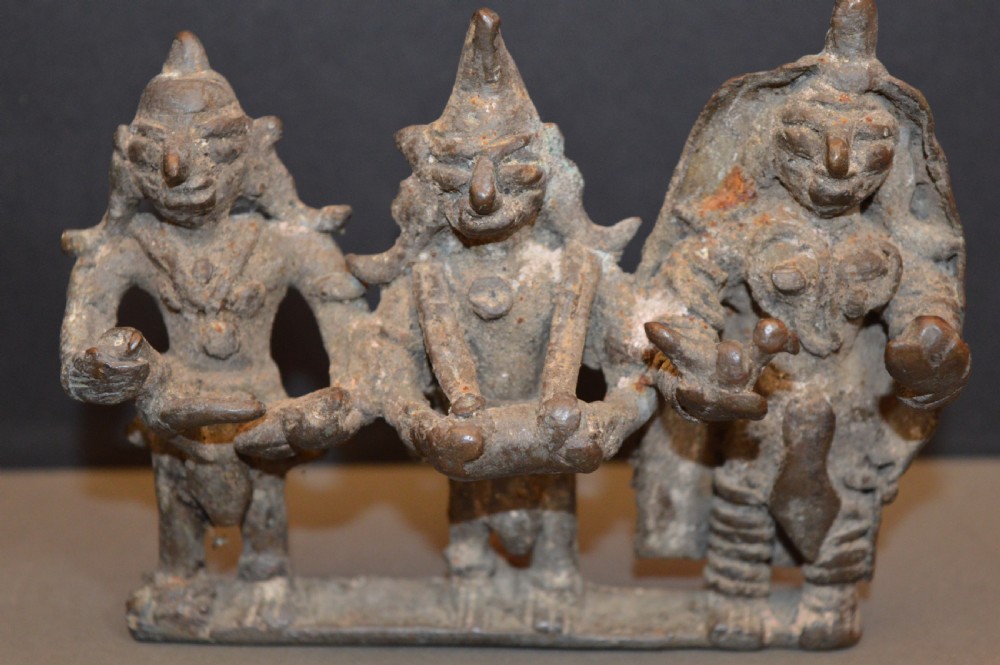 An antique dark bronze Indian sculpture featuring three standing Hindu deities with conical headwear on a shared rectangular base.