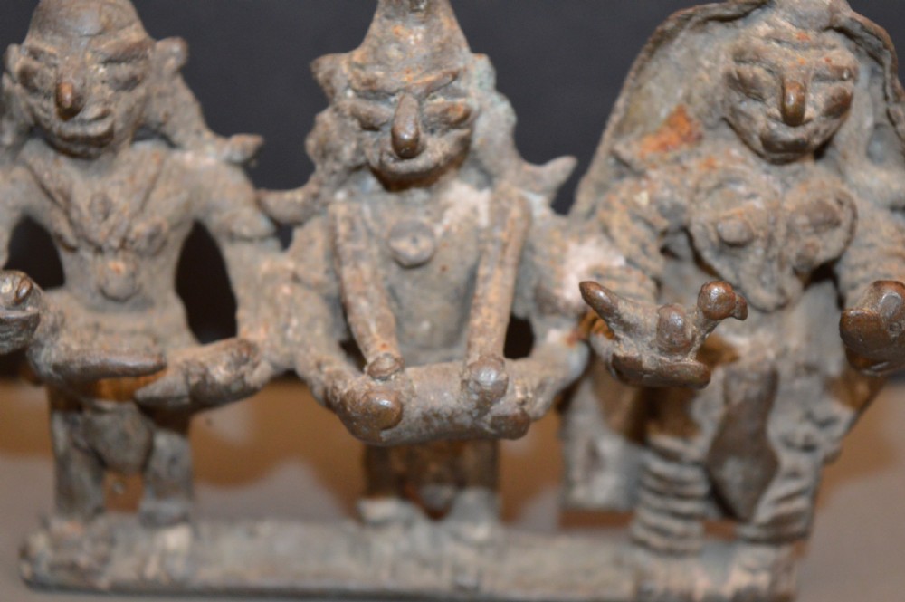 An antique dark bronze Indian sculpture featuring three standing Hindu deities with conical headwear on a shared rectangular base. View IV