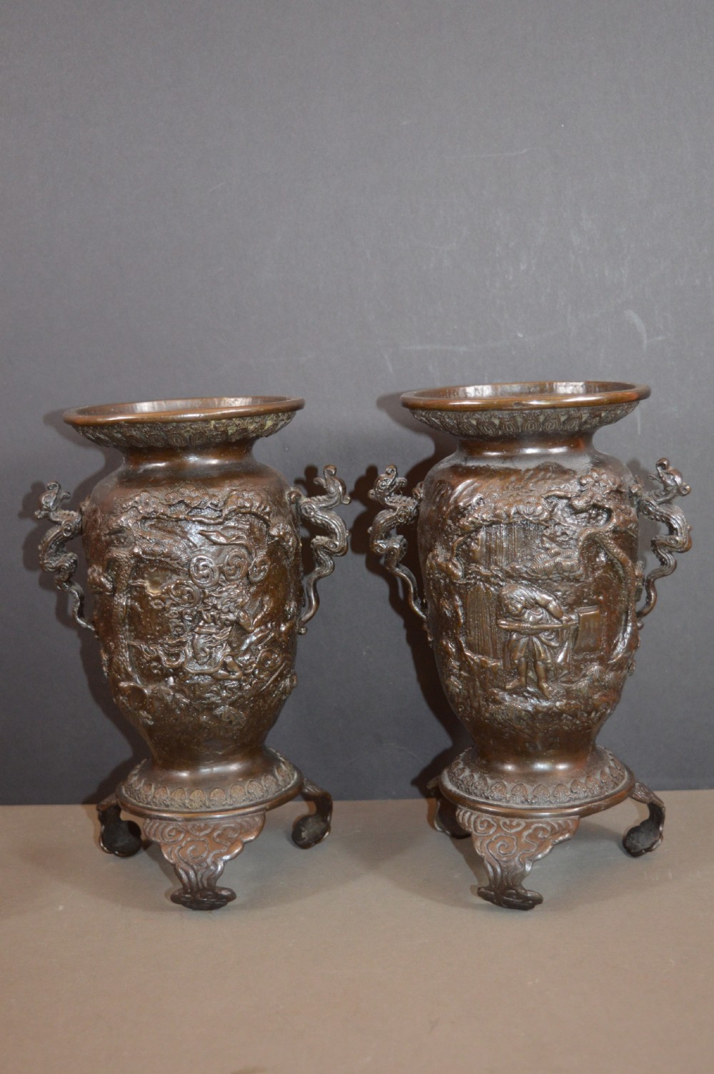 A pair of dark bronze Meiji period Japanese vases with high-relief figural decoration, dragon handles, and tripod bases.