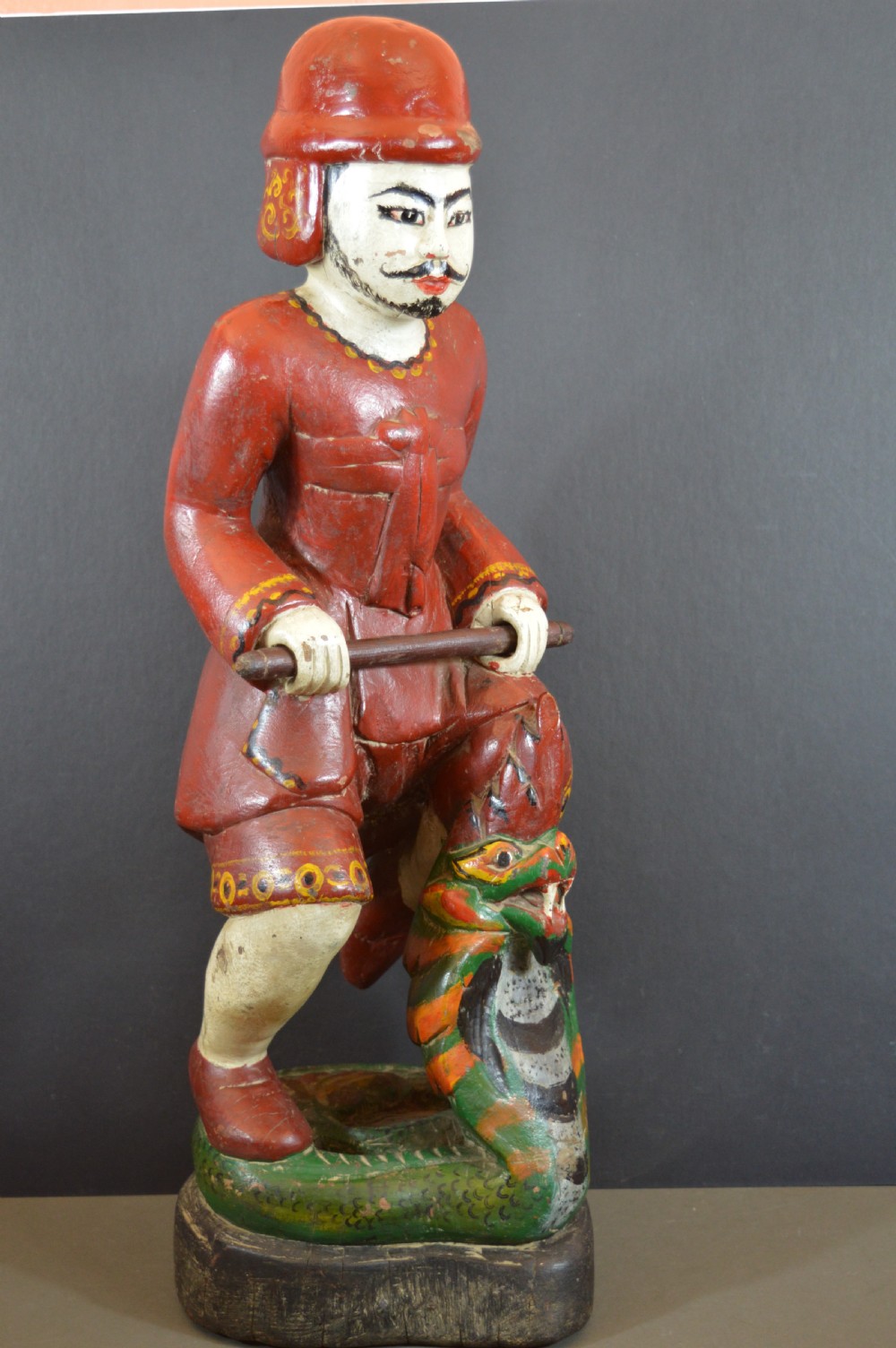 A late nineteenth-century carved wooden Tibetan figure painted in red, green, and white polychrome, depicting a standing male figure holding a rod above a serpent motif.