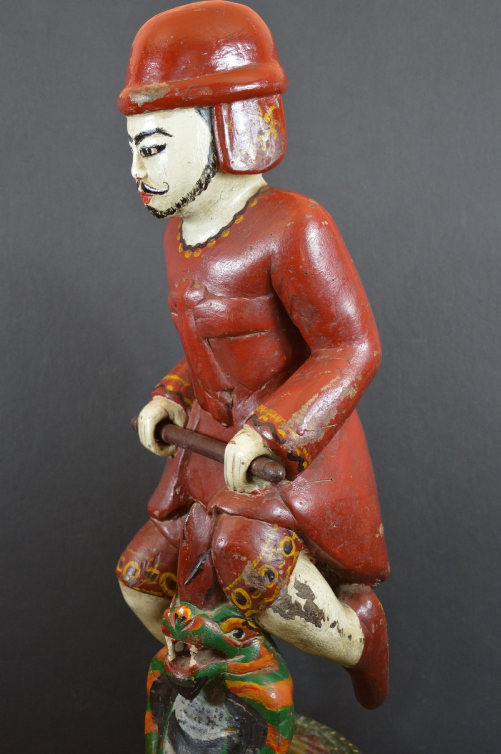 A late nineteenth-century carved wooden Tibetan figure painted in red, green, and white polychrome, depicting a standing male figure holding a rod above a serpent motif. View V