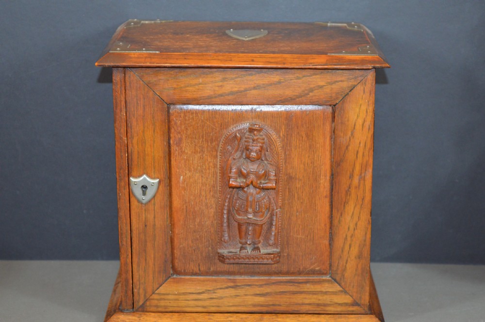 A late 19th-century oak desktop smokers cabinet featuring brass corner mounts and a central relief carving of an Indian deity.
