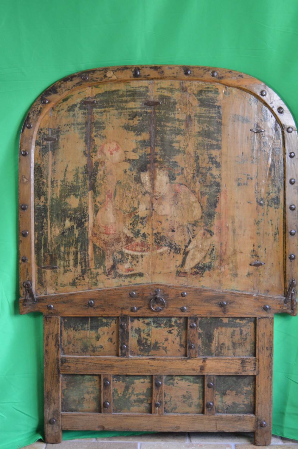 An arched antique Chinese wooden cart door featuring faded hand-painted figures, weathered green pigments, and iron studs.