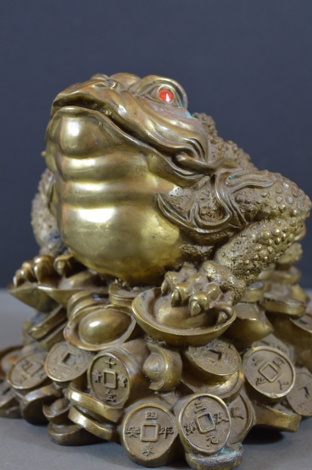 A heavy cast bronze three-legged money toad sculpture with red glass eyes, sitting on a base of Chinese coins and ingots.