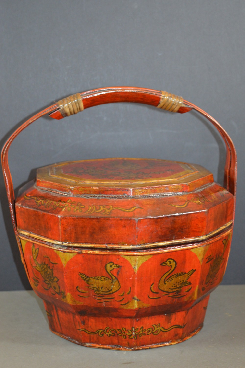 A red hand-painted Chinese wooden rice carrier featuring an arched overhead handle, lidded faceted body, and depictions of waterbirds and fish.