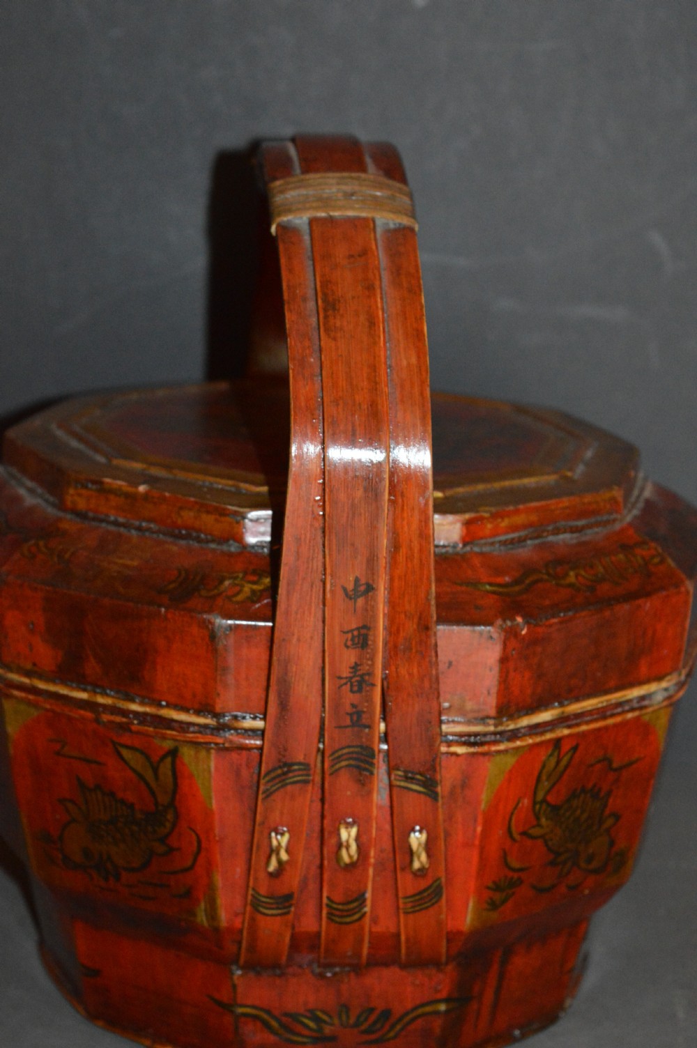 A red hand-painted Chinese wooden rice carrier featuring an arched overhead handle, lidded faceted body, and depictions of waterbirds and fish. View VI