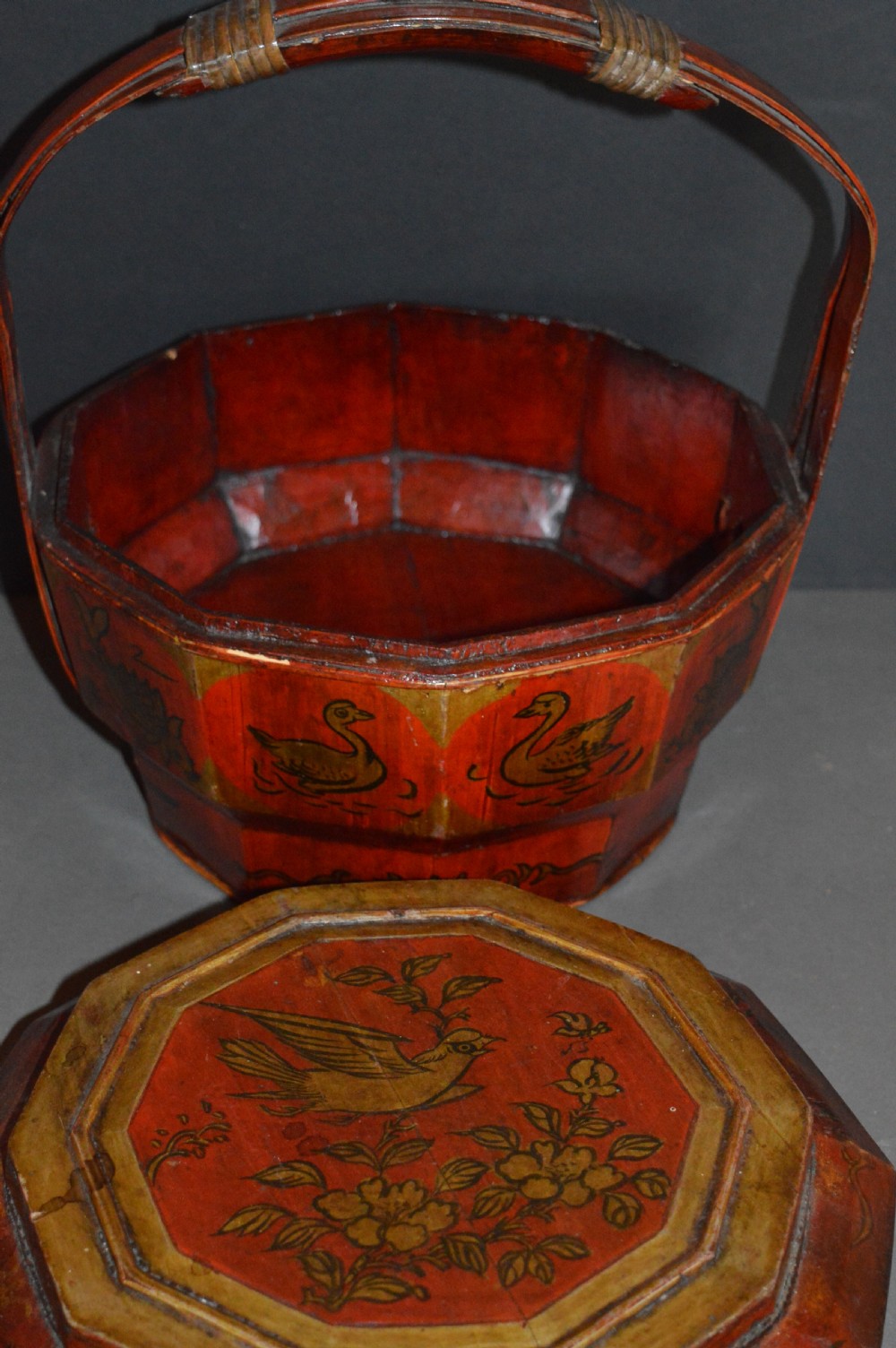 A red hand-painted Chinese wooden rice carrier featuring an arched overhead handle, lidded faceted body, and depictions of waterbirds and fish. View IX