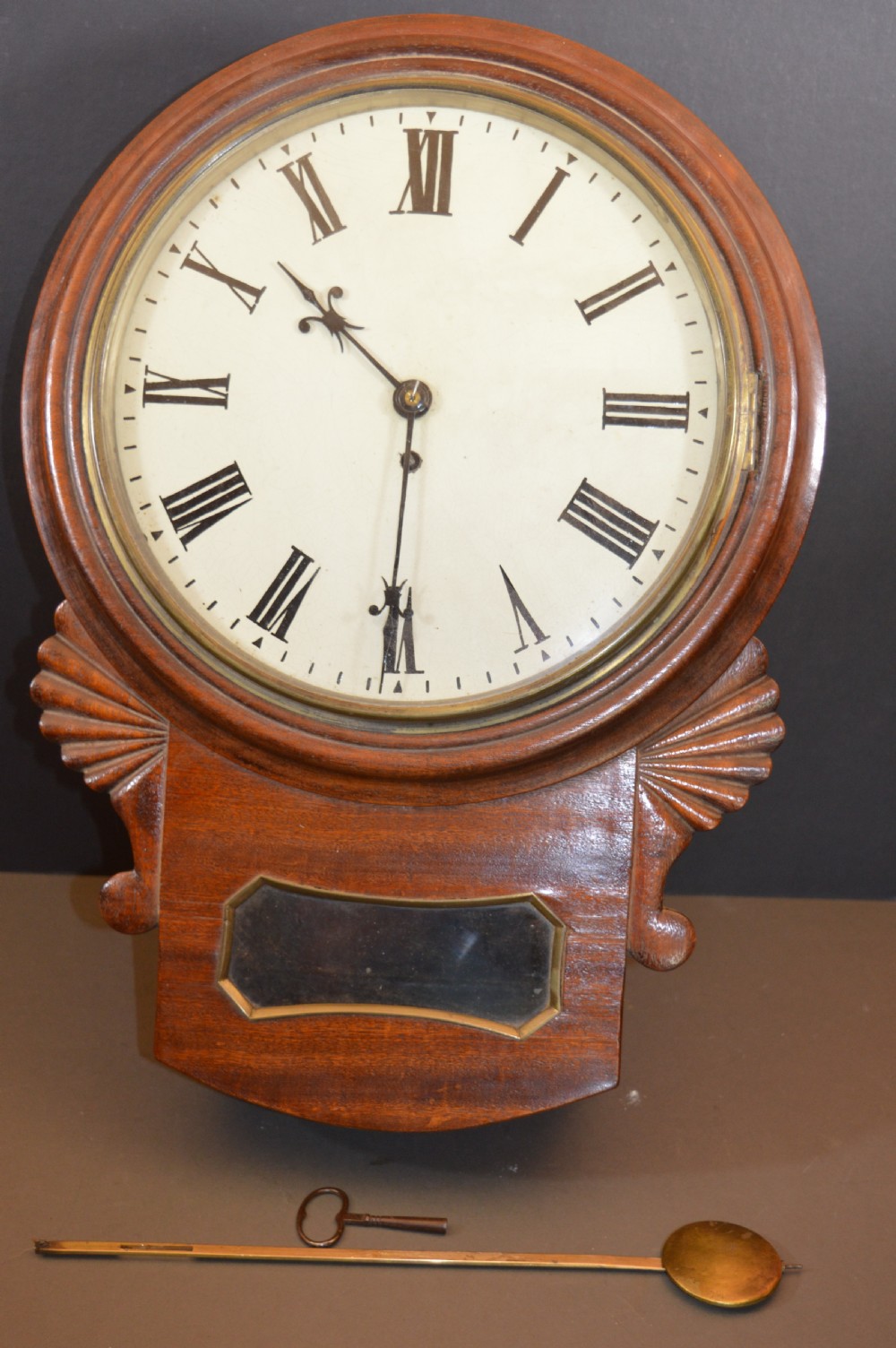 A Victorian mahogany drop dial fusee wall clock featuring a white circular dial with Roman numerals, carved side ears, and a lower pendulum window. View II