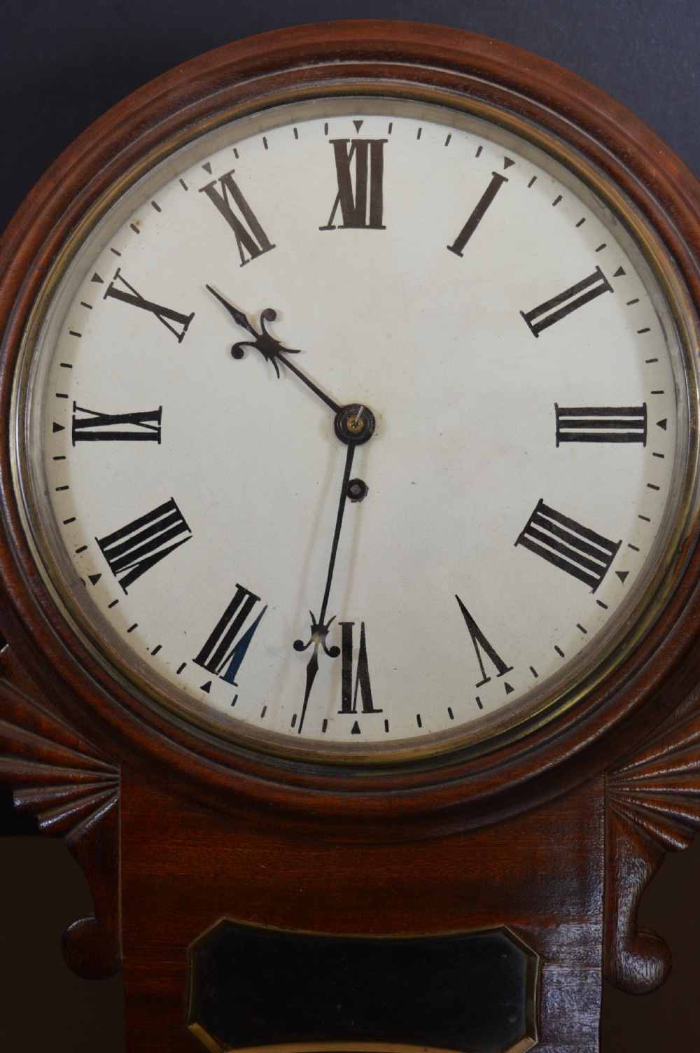 A Victorian mahogany drop dial fusee wall clock featuring a white circular dial with Roman numerals, carved side ears, and a lower pendulum window. View III