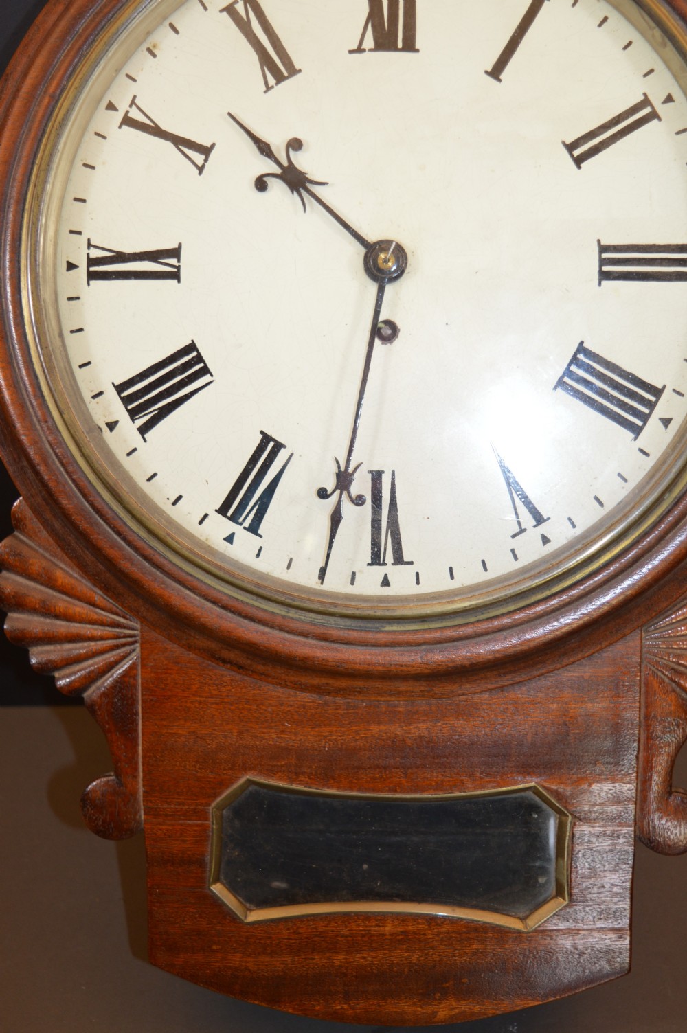 A Victorian mahogany drop dial fusee wall clock featuring a white circular dial with Roman numerals, carved side ears, and a lower pendulum window. View IV