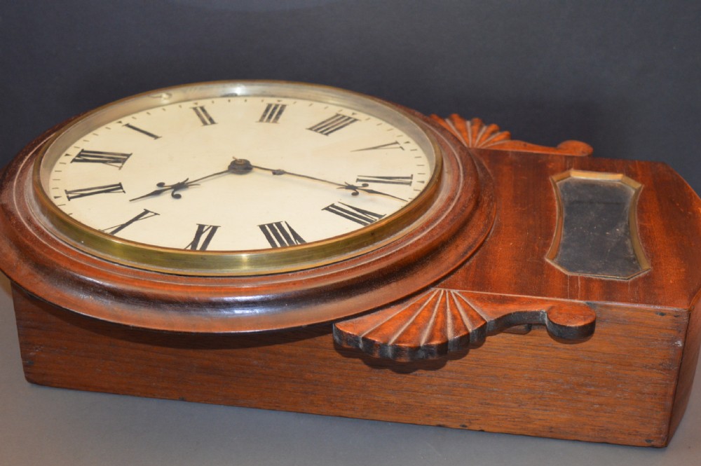 A Victorian mahogany drop dial fusee wall clock featuring a white circular dial with Roman numerals, carved side ears, and a lower pendulum window. View V