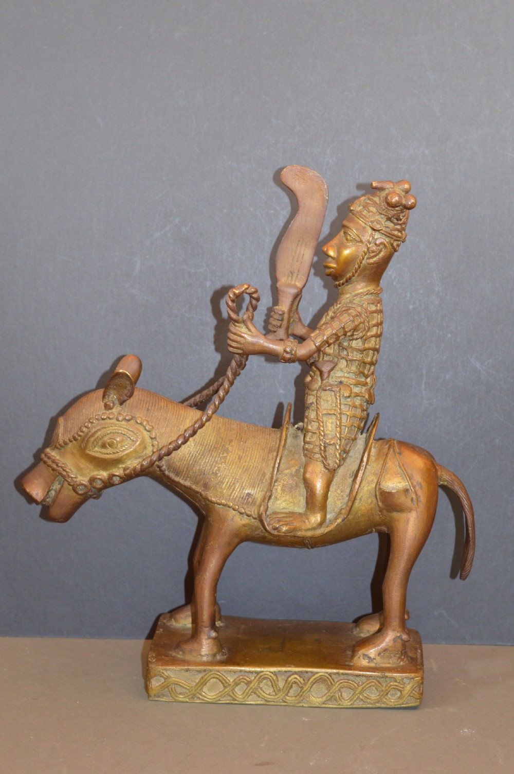 A cast metal Benin style sculpture of a warrior riding a decorated horse on a rectangular base, holding reins and a blade.