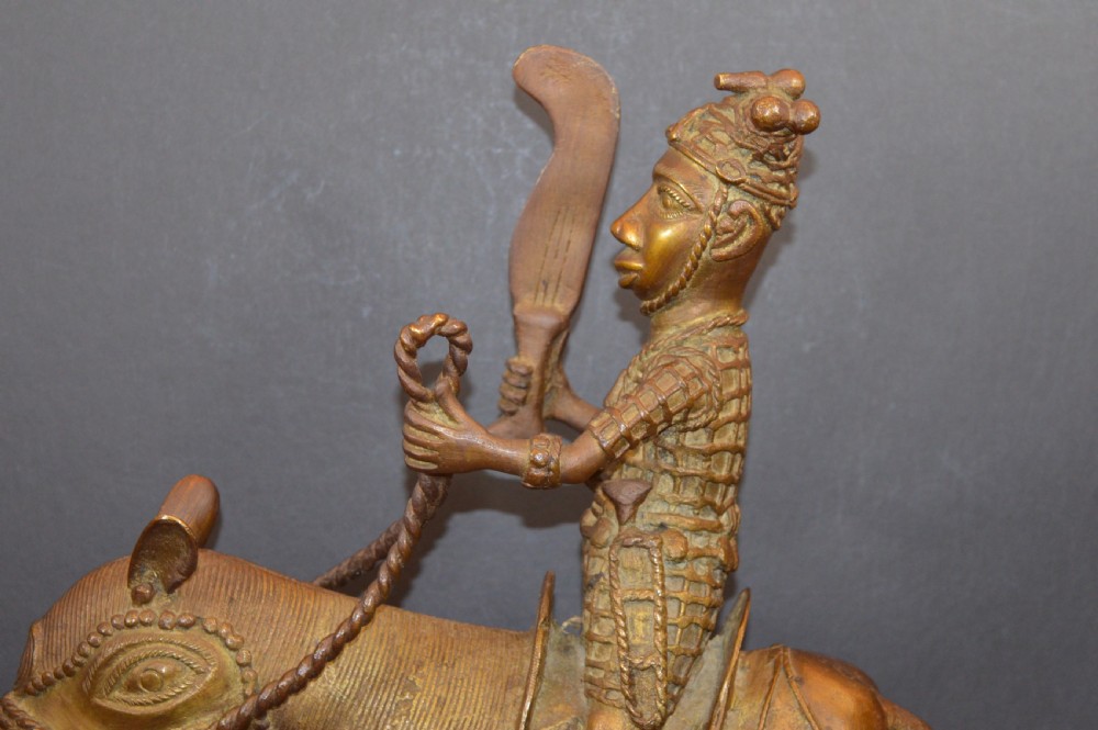 A cast metal Benin style sculpture of a warrior riding a decorated horse on a rectangular base, holding reins and a blade. View II