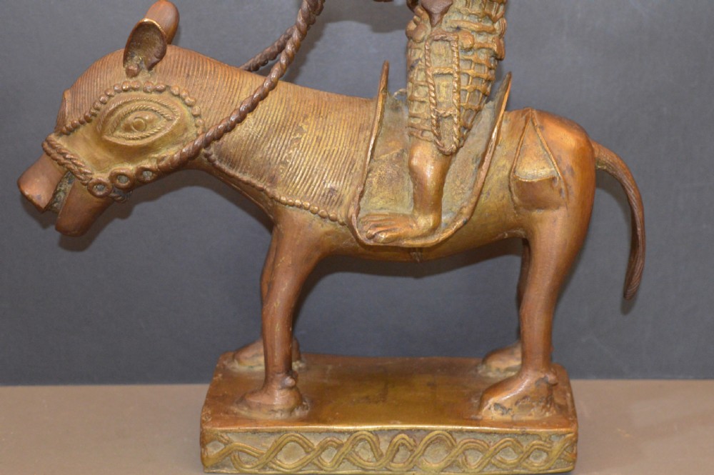 A cast metal Benin style sculpture of a warrior riding a decorated horse on a rectangular base, holding reins and a blade. View III