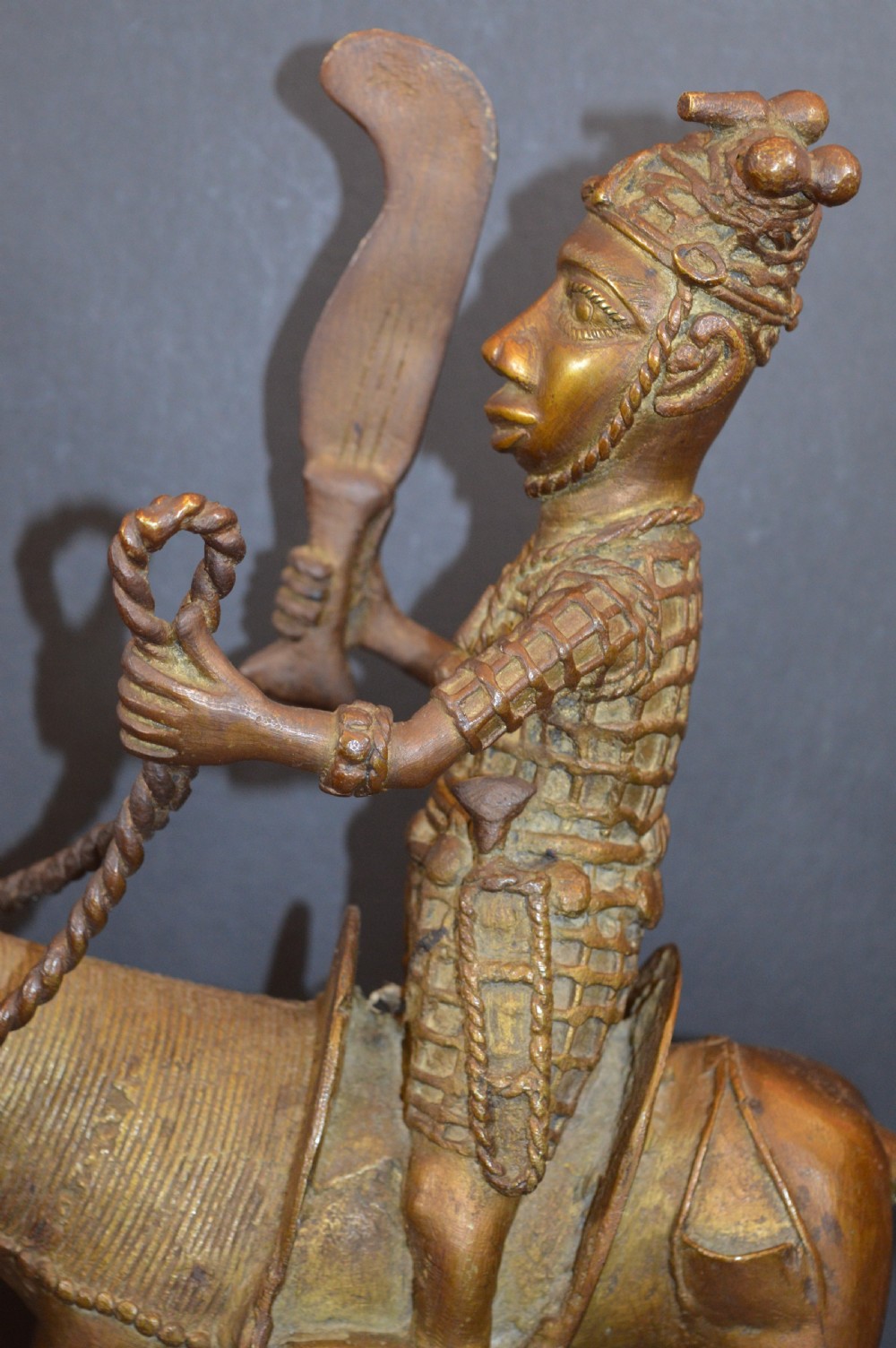 A cast metal Benin style sculpture of a warrior riding a decorated horse on a rectangular base, holding reins and a blade. View IV