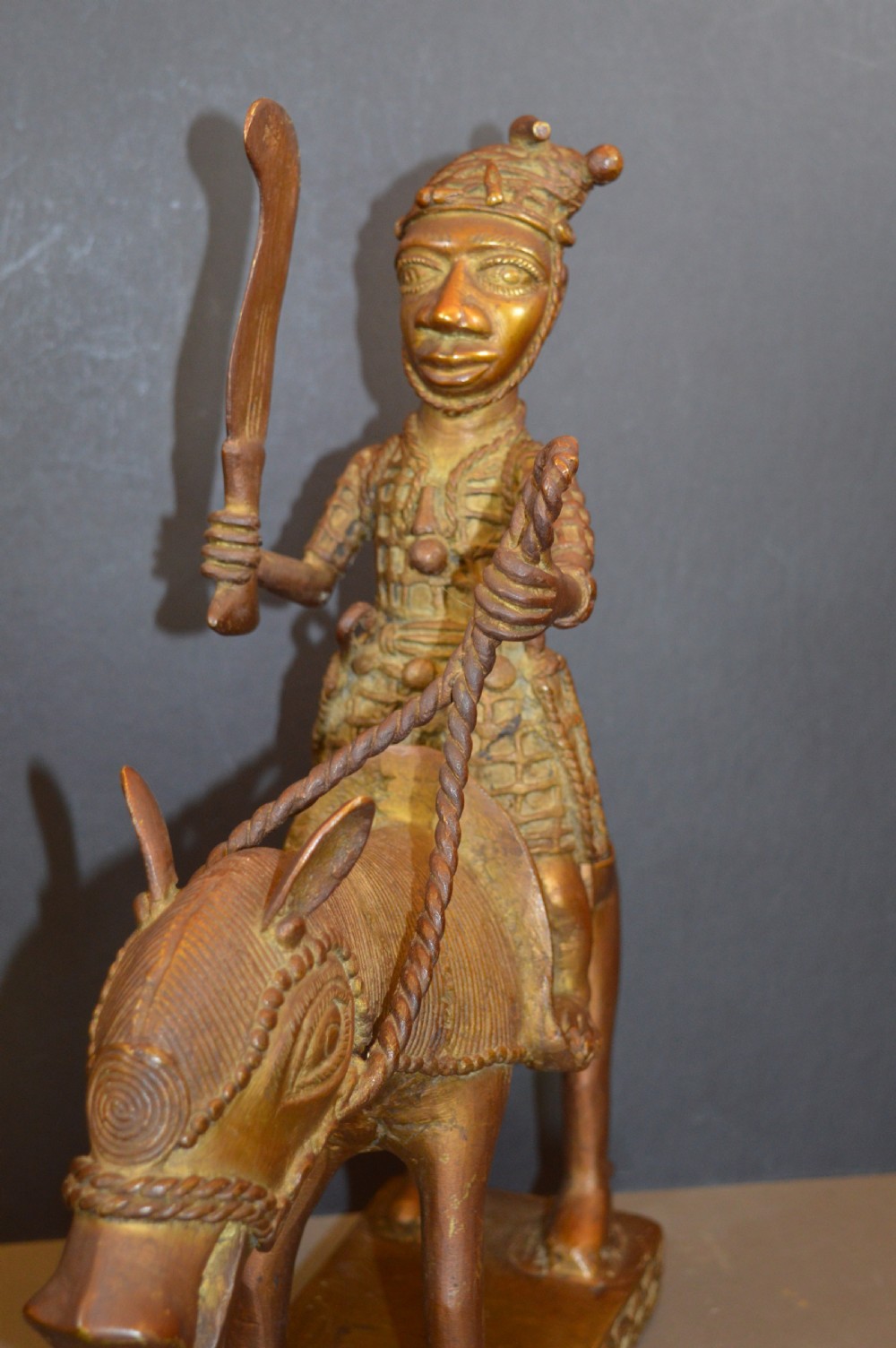 A cast metal Benin style sculpture of a warrior riding a decorated horse on a rectangular base, holding reins and a blade. View VI