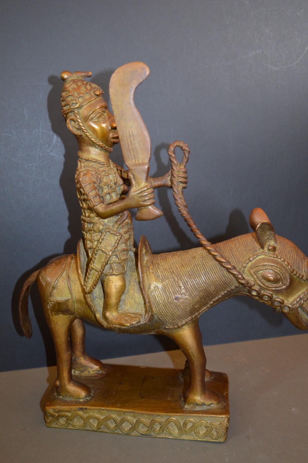 A cast metal Benin style sculpture of a warrior riding a decorated horse on a rectangular base, holding reins and a blade. View VIII