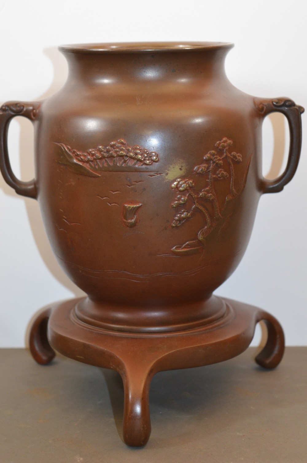 A Japanese Meiji period bronze urn featuring low relief carvings of pine trees and mountains with twin handles, resting on a matching carved bronze tripod stand.