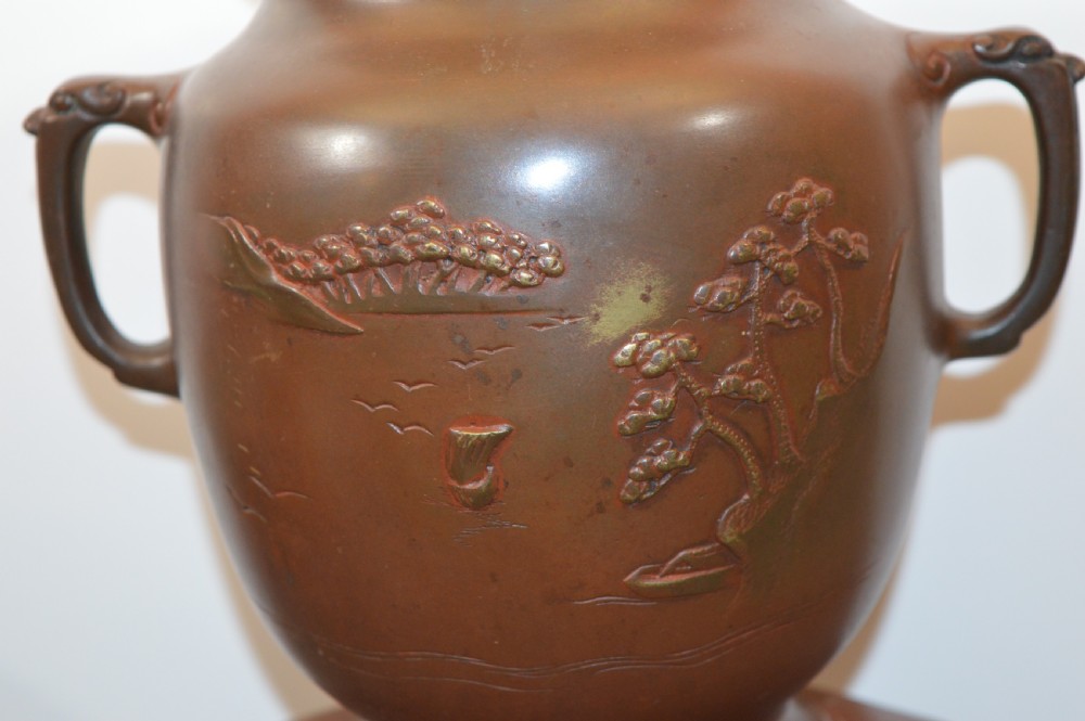A Japanese Meiji period bronze urn featuring low relief carvings of pine trees and mountains with twin handles, resting on a matching carved bronze tripod stand. View III