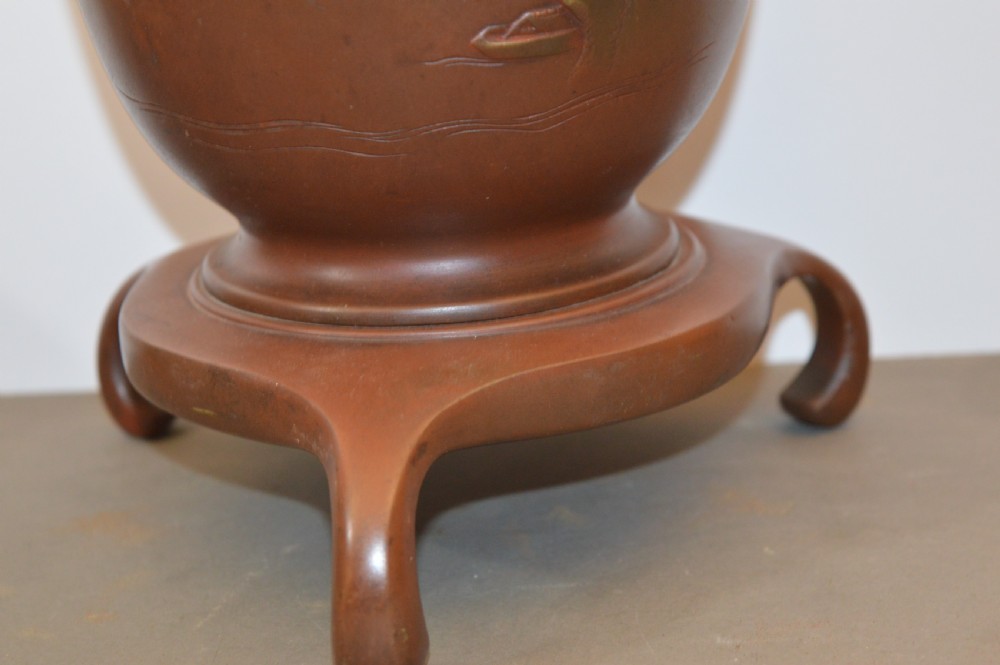 A Japanese Meiji period bronze urn featuring low relief carvings of pine trees and mountains with twin handles, resting on a matching carved bronze tripod stand. View IV